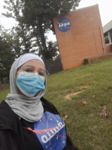 Joud wearing a cream colored hijab, a NASA shirt, and black sweater with a blue surgical mask. There is also the NASA brick building and trees behind her.