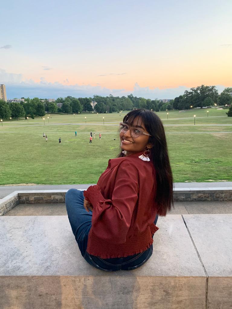 Shanika is seen sitting with a marroon colored jacket, blue jeans, glasses, long black hair, and smiling. She is turned around to smile. In the background, there is a field with people and a sunset.