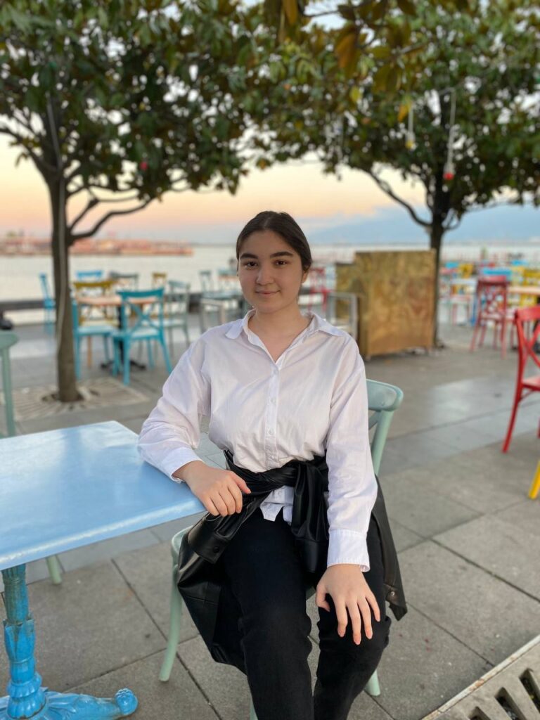 Farahnozkhon sitting at a table with a slight smile with trees and other chairs and tables behind her.