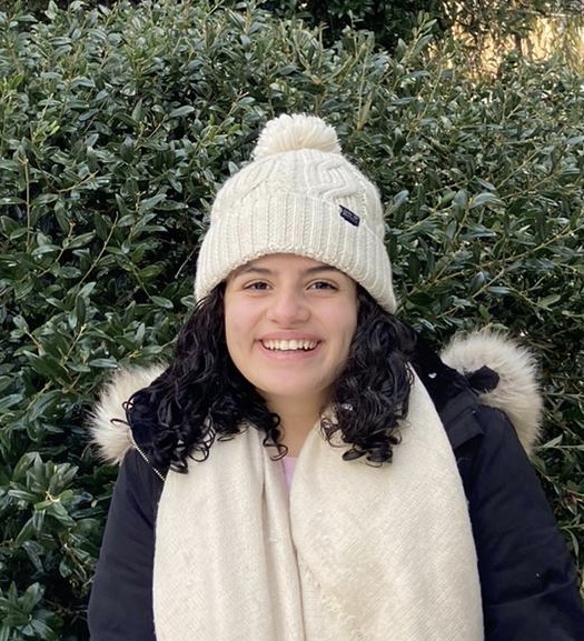 Leena is smiling in winter clothes while standing in front of greenery.