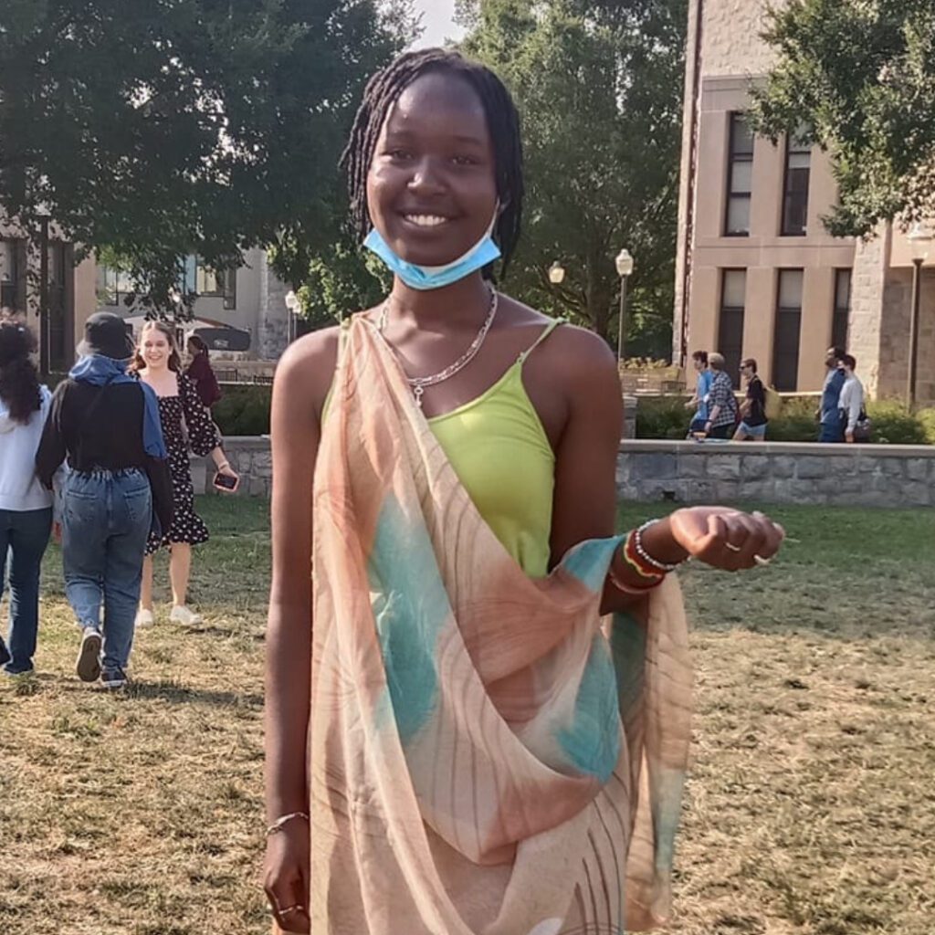 Elyse smiling on a grass lawn. There are people and buildings behind her.