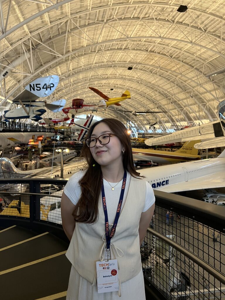 Young girl stands in center of image, smiling away from the camera. Background features a hanger with multiple large aircrafts