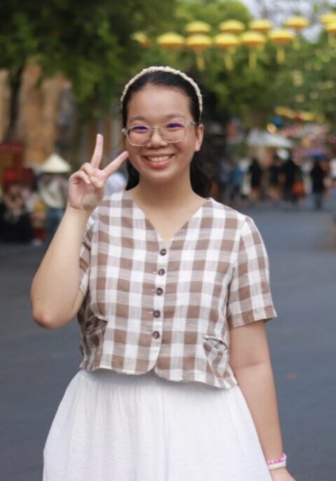 Ngan smiling in front of a blurry background. Her right hand is formed into the peace sign.